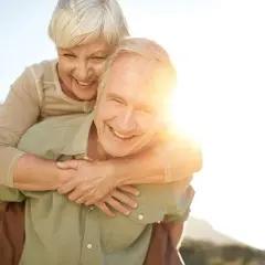 Ein Mann trägt eine Frau auf dem Rücken. Beide lachen und genießen die Sonne. Ein Bild von wahrer **Liebe im Alter**.