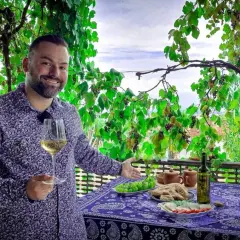 Mężczyzna z kieliszkiem białego wina, pod winoroślą, zaprasza do degustacji marani wino. Na stole owoce, chleb i ser.