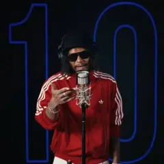 Raper w czerwonej bluzie z trzema paskami, w okularach i czapce, śpiewa do mikrofonu. Za nim neonowy napis "10". Jakby otulał go **łona kocyk** ciepła muzyki.