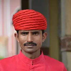Un homme porte un turban rouge vif et une chemise assortie. Sa moustache fine souligne son regard intense.