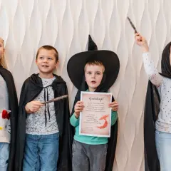 Cztery dzieci w strojach czarodziejów z różdżkami. Jeden chłopiec w kapeluszu czarownicy trzyma dyplom.