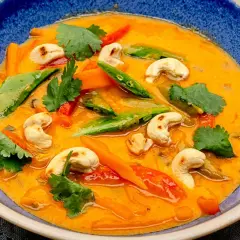 Ein cremiges rotes Thai Curry mit Cashewkernen, Zuckerschoten und Karotten in einer blauen Schüssel.