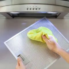 Hand reinigt mit gelbem Tuch den Metallfilter einer Dunstabzugshaube, um klebrige Rückstände zu entfernen.