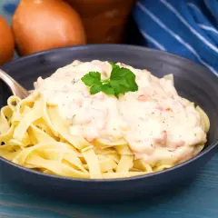 En skål med tagliatelle toppad med krämig ost och skinksås, garnerad med persilja. Två gula lökar syns i bakgrunden.