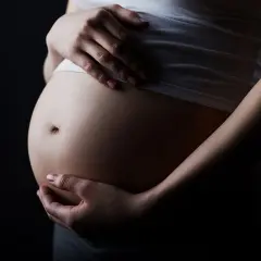 Brzuch kobiety w ciąży, z dłońmi delikatnie go obejmującymi, na ciemnym tle.