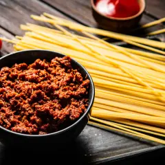Un tazón de carne boloñesa junto a espaguetis crudos y tomates. ¡Listo para unos deliciosos espaguetis a la boloñesa!