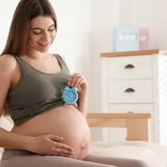 Uśmiechnięta ciężarna kobieta trzyma budzik nad brzuchem. Czas na poród zbliża się wielkimi krokami, czuć gorączkę przed porodem.