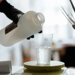 Ręka w czarnej rękawiczce przelewa przezroczysty płyn z butelki do plastikowego kubka stojącego na wadze.