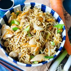 Bami Goreng mit Hühnchen, Karotten und Frühlingszwiebeln in einer Schüssel, daneben Sojasauce, Chili und frisches Gemüse.