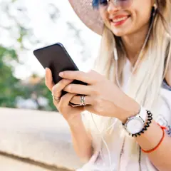 Uśmiechnięta kobieta w kapeluszu słucha muzyki przez słuchawki, trzymając telefon. Może szuka instrukcji, jak zainstalować radio w telefonie.