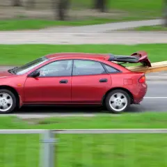 Czerwony sedan jedzie z otwartym bagażnikiem, w którym przewozi deski.