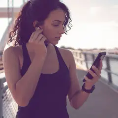 Frau auf einer Brücke, die sich Kopfhörer einsetzt und auf ihr Handy schaut. Sie ist bereit für ihre täglichen 10.000 Schritte.