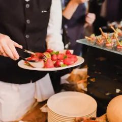 Kelner serwuje truskawki na talerzu, przygotowując elegancki **serwis francuski**. Obok widać przekąski na szklanej tacy.