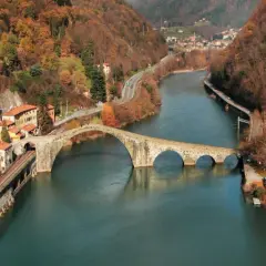 Il **Ponte del Diavolo**, con le sue arcate in pietra, attraversa un fiume circondato da colline autunnali e case.