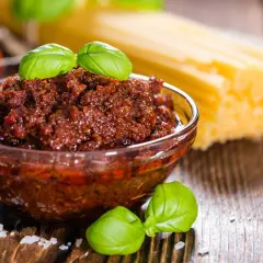 Pesto di pomodori secchi in una ciotola di vetro, con spaghetti e basilico sullo sfondo.