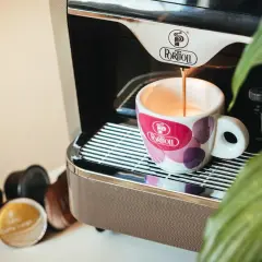 Espresso pouring into a patterned cup next to coffee capsules and a coffee machine.