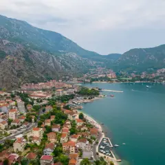 Widok na zatokę i miasto Kotor w Czarnogórze. Domy z czerwonymi dachami schodzą ku turkusowej wodzie, a nad wszystkim górują strome, skaliste wzgórza.