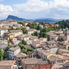 Panoramica di un borgo collinare con case dai tetti rossi, una chiesa con campanile e montagne sullo sfondo. Un paesaggio incantevole nei dintorni di Rimini.