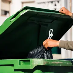 Ręka wrzuca worek ze śmieciami do zielonego kontenera. Zastanawiasz się, ile wynosi opłata za śmieci?