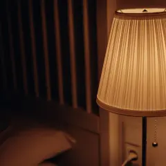 Lámpara con pantalla cilindrica de tela plisada, iluminando suavemente una habitación oscura con una cama al fondo.