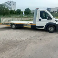 Biała laweta z platformą stoi na parkingu przed budynkami.