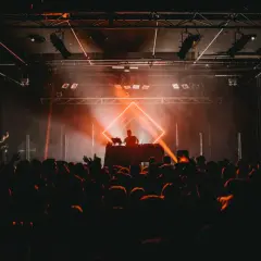 DJ set sous un éclairage orange vif, ambiance techno année 90. Foule en délire devant la scène.