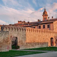 Antica cinta muraria in pietra delle Crete Senesi, con torri medievali e cielo al tramonto.