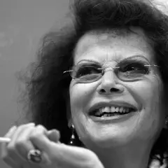 Uśmiechnięta Claudia Cardinale, ikona kina, z papierosem w dłoni. Jej spojrzenie pełne jest życia, jak w jej najlepszych filmach.