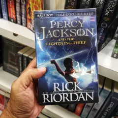 Dłoń trzyma okładkę książki "Percy Jackson i Złodziej Pioruna". Na okładce młody bohater z mieczem w ręku, otoczony błyskawicami.