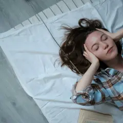 Junge Frau mit geschlossenen Augen und Händen an den Schläfen auf einem Bett liegend. Ein Buch liegt daneben. Tipps für gesunden Schlaf.