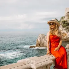 Kobieta w czerwonej sukience i kapeluszu siedzi na balustradzie z widokiem na morze. Idealny strój, co zabrać na plażę w Chorwacji.