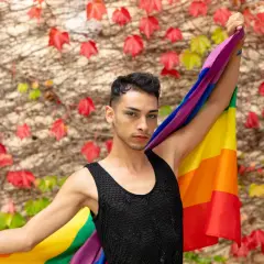 Un giovane con la bandiera arcobaleno, simbolo del **queer significato** di libertà e identità, si staglia contro un muro ricoperto di foglie rosse e verdi.