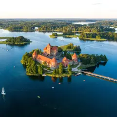Zamek Troki, malowniczo położony na wyspie pośród jeziora, otoczony zielenią i wodą, z mostem prowadzącym do jego bram.