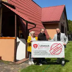 Trzy osoby trzymają baner "BUDYNEK ZALEGALIZOWANY" od Legasus. Wzór zgłoszenia samowoli budowlanej jest widoczny.
