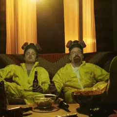 Die Besetzung von Breaking Bad, Walter und Jesse, entspannt in ihren Schutzanzügen mit Bier und Snacks auf dem Sofa.