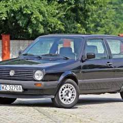 Czarny VW Golf 2, klasyk z lat 80., lśni w słońcu na tle ceglanego muru i zieleni.