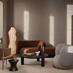 Ein stilvolles Wohnzimmer mit einer rostbraunen Couch. Ein beiger Bouclé-Sessel und ein Holzstuhl ergänzen die Sitzgelegenheiten. Ein runder Couchtisch mit Deko-Objekten steht im Zentrum.