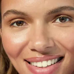 Una mujer sonríe, mostrando su piel radiante. Sostiene un producto con agua de rosas, ideal para la doble limpieza facial.
