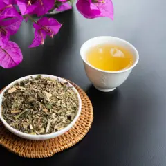 Una tazza di tisana per digerire accanto a una ciotola di erbe secche, con fiori viola sullo sfondo.