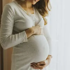 Kobieta w ciąży, z dłońmi na brzuchu, wygląda na szczęśliwą. Jej uśmiech sugeruje, że jest pełna życia, jak nimfomanka.