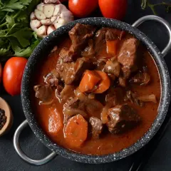 Gulasz z sarny w głębokim sosie, z marchewką i cebulą, obok przypraw i pomidorów.