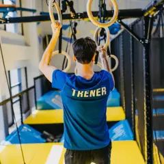 Trener w niebieskiej koszulce z napisem "TRENER" trzyma się kółek gimnastycznych. W tle widać trampoliny i siatki. To miejsce, gdzie można wzmocnić powięź.