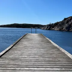 Träbrygga leder ut mot det glittrande vattnet vid Grebbestad strand. Klippor och öar i bakgrunden under en klarblå himmel.