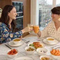 Deux femmes trinquant avec des jus verts et orangés, entourées d'un repas sain. Un exemple de comment ne pas grossir à la ménopause.