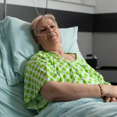 Starsza kobieta w szpitalnym łóżku, z tlenem przy nosie, w zielono-białej bluzce w groszki. Czeka na leki, być może Duracef.
