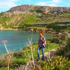 Malta w marcu: kobieta na ścieżce z widokiem na turkusową zatokę i zielone wzgórza. Pogoda idealna na spacer.