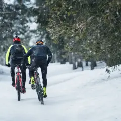 Dwóch rowerzystów przemierza zimowe trasy rowerowe Olsztyn, podczas gdy śnieg delikatnie opada na las.
