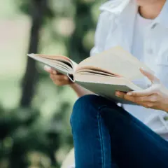 Osoba w dżinsach i białej kurtce czyta książkę na łonie natury. Wśród zieleni, niczym pingwiny czytelnicy, zanurza się w lekturze.