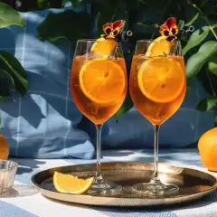 Dwa Aperol Spritz z plastrami pomarańczy i kwiatami, otoczone pomarańczami i liśćmi. Idealne aperol spritz składniki na letni dzień.