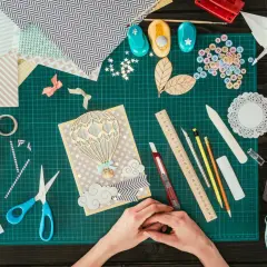 Ręce tworzą kartkę z balonem na gorące powietrze. Wokół materiały do scrapbookingu, taśma, nożyczki, guziki i inne akcesoria.